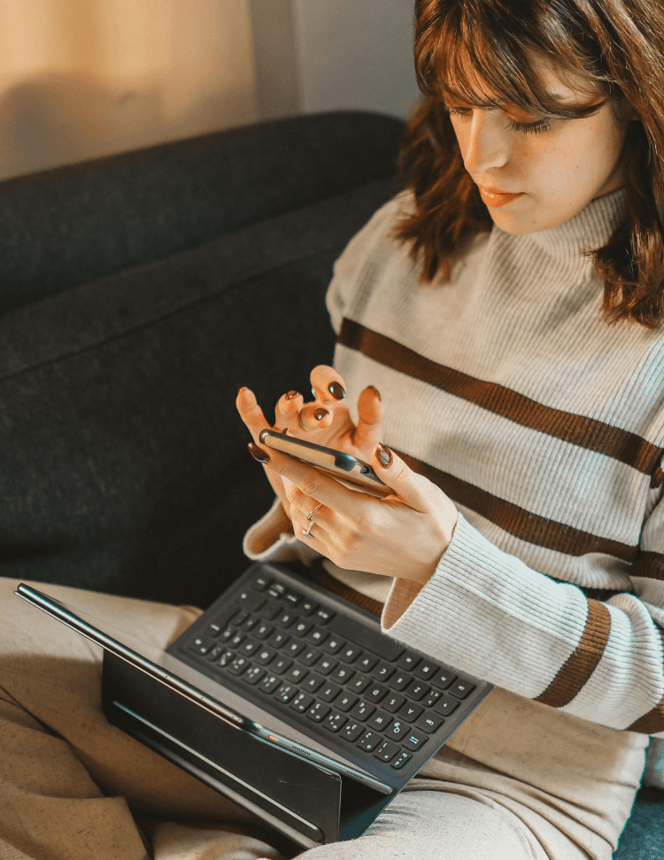 Woman sitting on a sofa with a laptop on her lap, while she is texting on her phone.