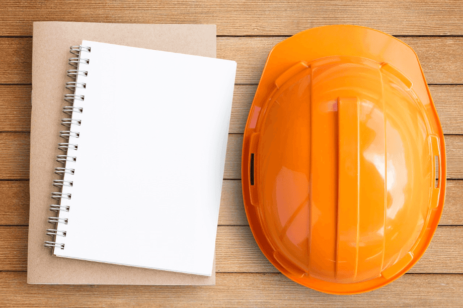 An hard hat placed on a wooden surface next to a spiral notebook and a folder, symbolizing safety and planning.