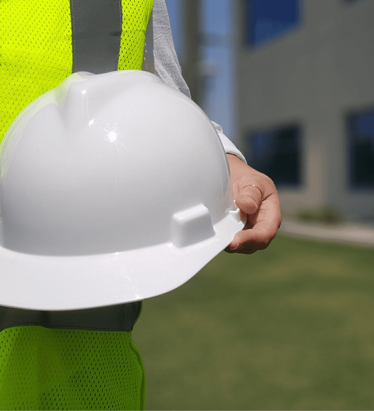 A person wearing a high-visibility vest holding a white hard hat outdoors, emphasizing safety and professionalism.