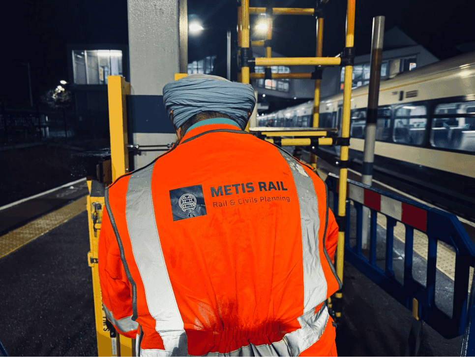 A Metis Rail worker wearing high-visibility safety gear at a rail station worksite at night, with scaffolding and a train in the background.