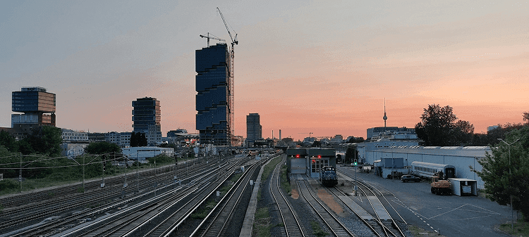 Railway tracks leading toward modern high-rise buildings under construction, with a train yard and sunset sky in the background.